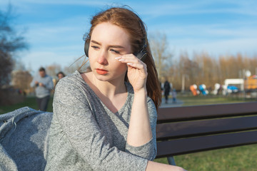 Young, sad cry redhead girl in the spring in the park near the river listens to music through wireless bluetooth headphones