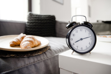 An alarm clock is standing on the coffee table. A pair of croissants is laying in the plate on the...