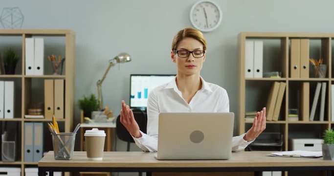 Charming young woman in the white shirt and glasses sitting at the office table with laptop and meditating. Indoor