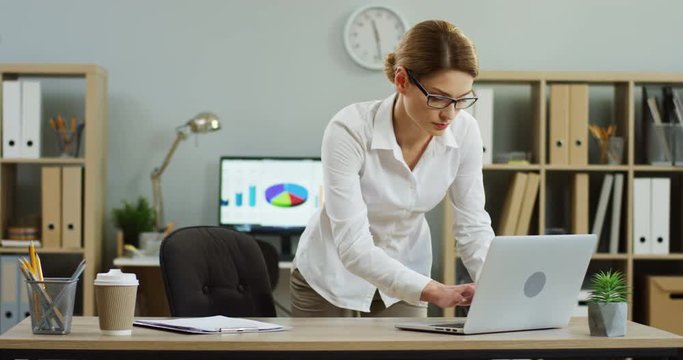 Caucasian good looking female office worker entering some data on the laptop computer from a document while standing near the workplace in the modern office. Indoors.