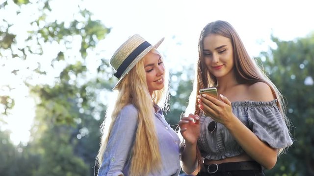 Happy pretty girls using a smartphone relaxing in the park at beautiful sunlight on warm summer day.