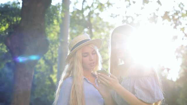 Two attractive young women smiling while looking at smartphone standing on the street of modern city in sunlight.