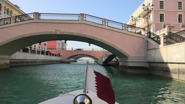 Venetian bridge on canals of picturesque Qanat Quartier icon of Doha, Qatar from a touristic boat with flag of Qatar. Venice at the Pearl, Persian Gulf, Middle East. Famous attraction at sunset light.