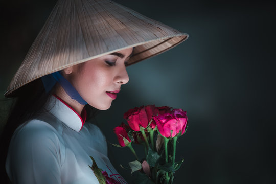 close up Vietnamese gill in white dress wearing a hat and holding red rose in hands on dark background