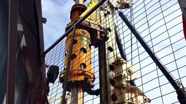 Wide angle looking upwards through fence at a hydraulic rotary drilling rig as it is raised and twirled. on a construction site.