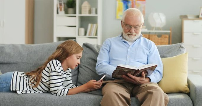 Grey haired old grandfather in the glasses reading a book and his granddaughter using smartphone while they resting on the couch.