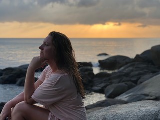 Fototapeta na wymiar a light-skinned brown-haired girl in light beach clothes sits on the stones of the coastline of Karon beach in Phuket in Thailand and admires the late sunset
