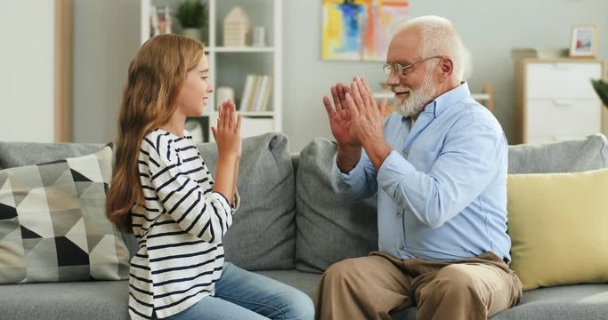 Caucasian cheerful grandfather and granddaughter sitting on the couch in the living room and playing hands game.