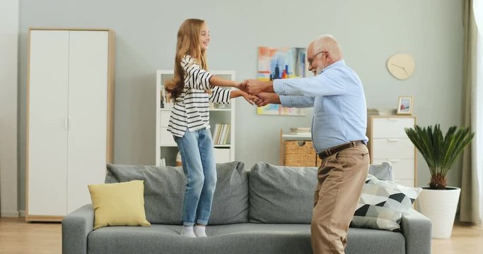 Happy cute teen girl jumping on the couch in the living room with her old grandfather.