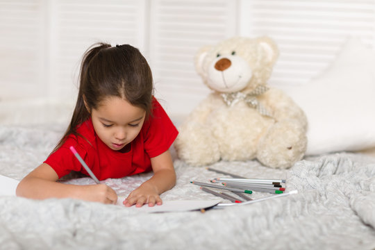 cute little child girl in red pajamas with teddy bear draws with pencils at home
