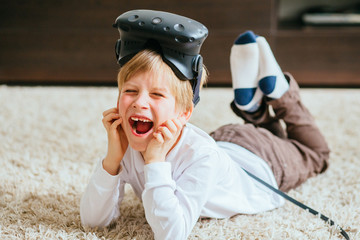 Fototapeta premium Excited blond boy wearing virtual reality goggles lying on floor using virtual reality headset and gamepads in the light living room at home.
