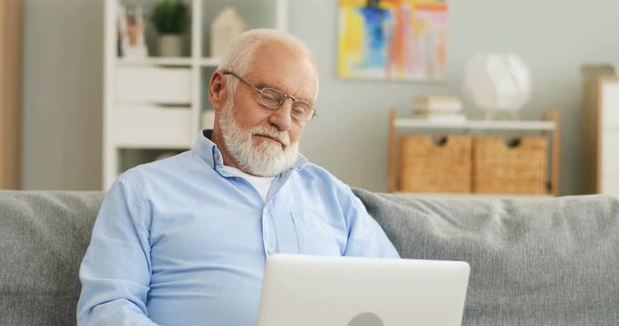 Senior man with grey hair and in glasses sitting on the couch, watching something on the laptop computer and smiling. Portrait.