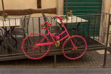 An old fashioned cycle, painted pink and leaning against the railings of a restaurant and with the frame and wheels decorated by flowers