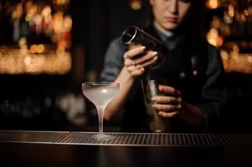 Female bartender pouring a cocktail in steel shaker on the foreground of ice glass