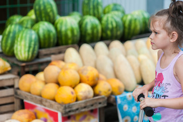 Teenager girl on a scooter stands near the counter with watermelons and melons