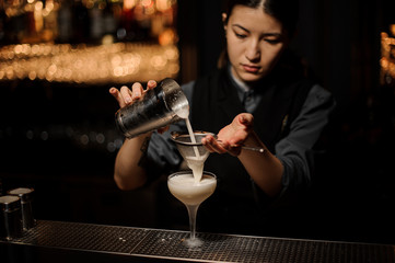 Bartender girl pouring a delicious cocktail from the steel shaker through the sieve