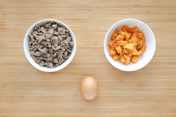 Chopped of boiled pork liver and sweet potato in white bowls on wooden board with egg like an owl. Raw healthy baby food.