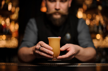 Bartender with a beard serving a cocktail in the glass with foam decorated with a one red rose bud