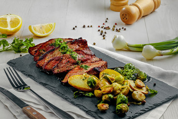 Delicious pan-seared steak with vegetables on a stone board. Meat meal with brussels sprouts, potatoes, onions and herb butter on a white wooden table.