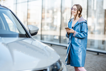 Lifestyle portrait of a young stylish woman in blue coat using phone near the modern car outdoors