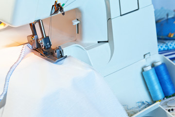 Processing the edges of white fabric with a finishing seam with blue threads on an overlock machine. Sewing clothes at home. Household. Close-up