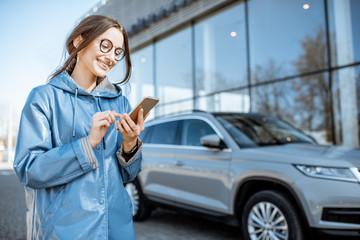 Lifestyle portrait of a young stylish woman in blue coat using smart phone near the modern car outdoors