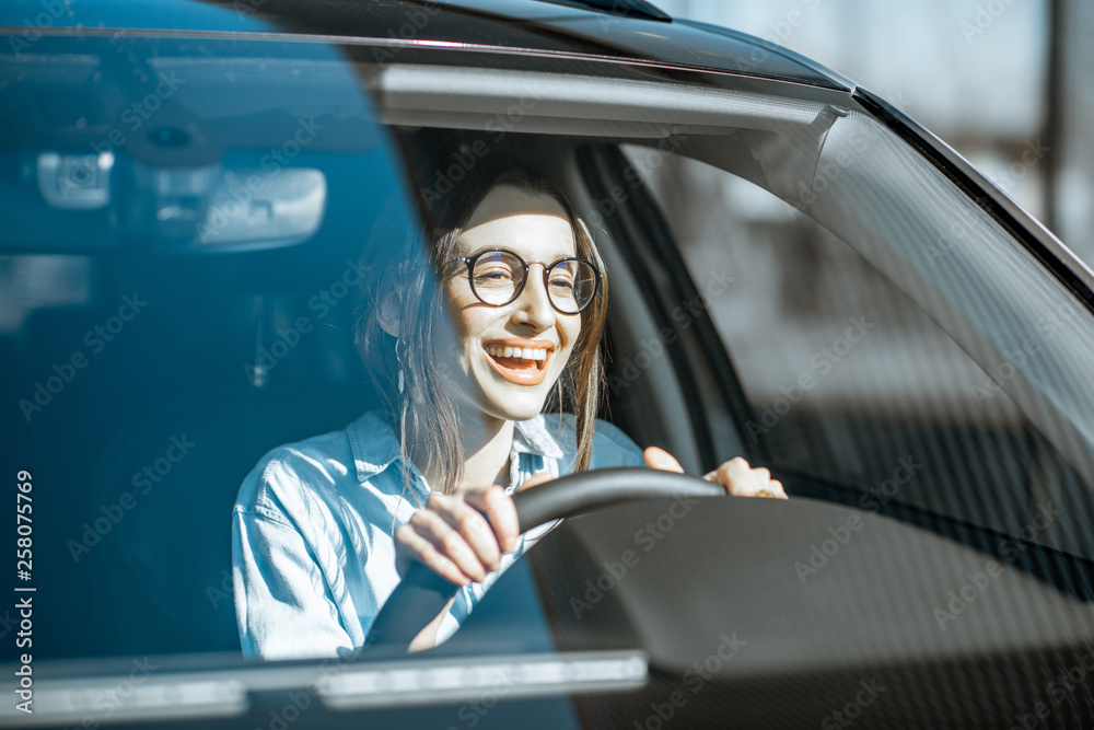 Wall mural Young and happy woman driving a luxury car, front view through the windshield with sunlight