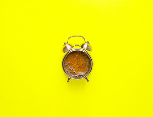 Alarm clock with freshly brewed black coffee with bubbly crema instead of clock face in the middle on bright yellow background. Flat lay. Morning sunlight breakfast energy. Creative food art