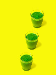 Row of three glasses of leafy greens smoothies spinach vegetables fruits on bright sunny yellow background. Healthy plant based diet detox vegan concept. Creative food poster. Vertical