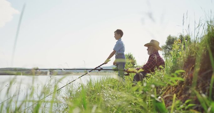 Old grandfather in a hat fishing together with his grandson on the beautiful lake. Outdoor.