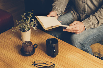 Hombre leyendo un libro con una taza de café sobre una mesa de madera rústica. Vista superior - obrazy, fototapety, plakaty