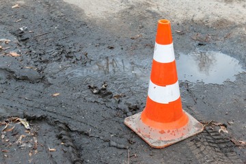 close-up of striped plastic orange parking cone on wet ground with car tracks, puddles and orange leaves. Warning orange cone on the сonstruction site with copy space background, selective focus.