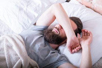 Monday morning again. Laziness. Man lying in bed covering eyes with arm against daylight.