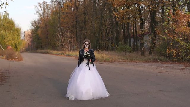 A young girl with a creepy make-up in the form of a skull on her face in a wedding dress and a leather jacket going on an empty road. Halloween. The image of the dead bride.