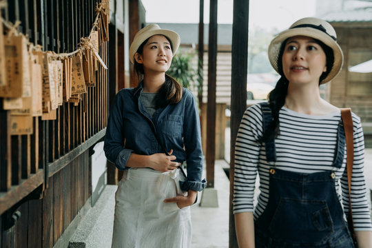 two asian female travelers join tour guided in japanese local traditional shrine house temple in garden corridor in tokyo. young women with hats walk along wish wooden wall curious look around relax.