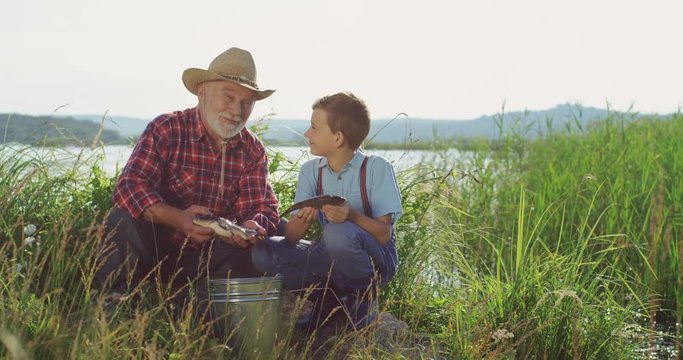 Portrait of the senior grandfather and grandson sitting in the grass on the shore of the lake with fish in hands and smiling to the camera. Outside.