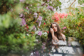 Young beautiful caucasian woman in glass greenhouse among colorful azalea flowers. Art portrait of a long-haired girl wearing a romantic red dress.