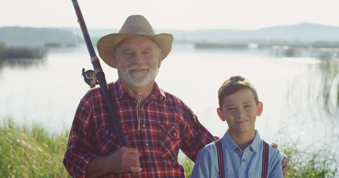Portrait of the old gray haired fisherman with gis grandson standing at the lake shore, looking at each other and then smiling to the camera. Outdoor.