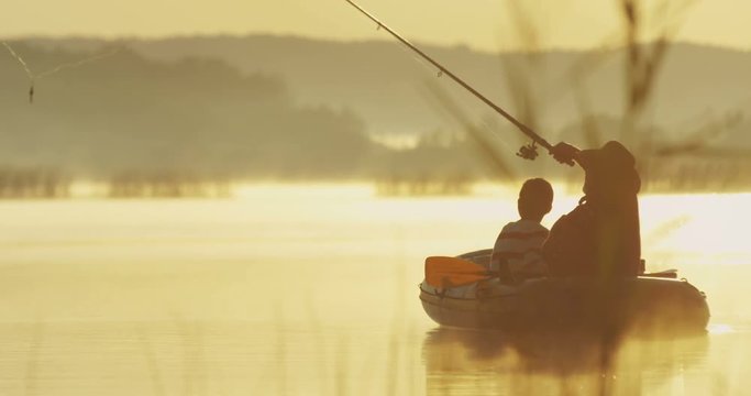Senior fisherman in a hat with his grandson sitting in the boat in the cane on the lake at the dawn and throwing a rod in the water. Outdoors.