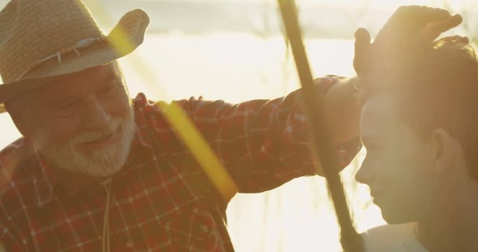 Close up of the senior grandfather in a hat caresing his grandson on the head while they fishing in the cane at the dawn. Outdoor.