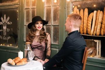 Beautiful young, stylish pregnant girl with her husband sitting at a table with croissants on the background of a French bakery