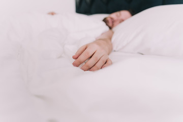 The hand of a man rests on white sheets. The man sleeps in a large bed of a hotel. It is located in...