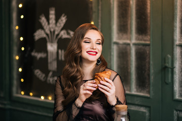 Portrait of a beautiful brunette with curls and red lipstick with a croissant in her hands on the background of a French bakery