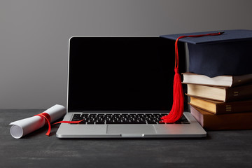 Laptop with blank screen, diploma, books and academic cap with red tassel isolated on grey