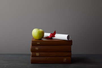 Brown books, diploma with ribbon and green apple on textured surface on grey