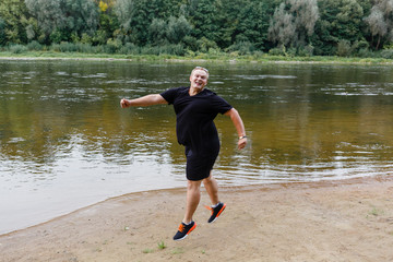 very fat guy jumps on the river bank in the park in summer