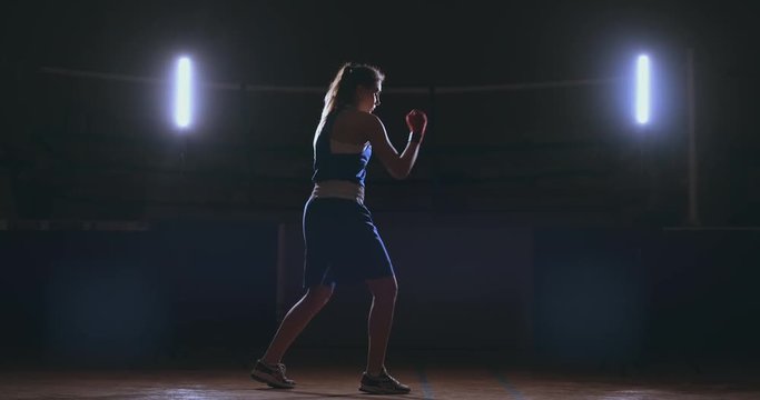 A beautiful woman boxer trains in a dark gym and works out punches in slow motion. side view. Steadicam shot