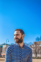 Portrait young bearded guy with emotions in shirt on the background of the blue clear sky in the park. Street fashion, hipster.