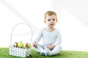 cute baby sitting near straw basket with Easter eggs isolated isolated on white