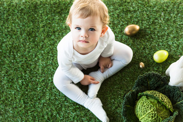 top view of adorable child looking at camera while sitting near Easter eggs, decorative rabbit and savoy cabbage